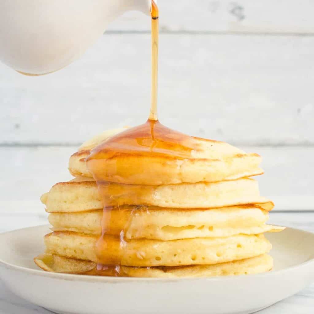 White syrup cup pouring maple syrup over stack of pancakes.White syrup cup pouring maple syrup over stack of pancakes.