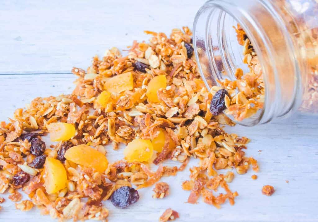 Clear mason jar on its side with fruit and nut granola spilling out onto white wood counter