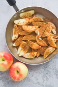 Apple slices sprinkled with cinnamon in a skillet alongside two whole apples on counter