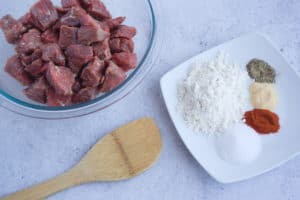 Clear bowl of raw stew meat next to white plate of flour and spices