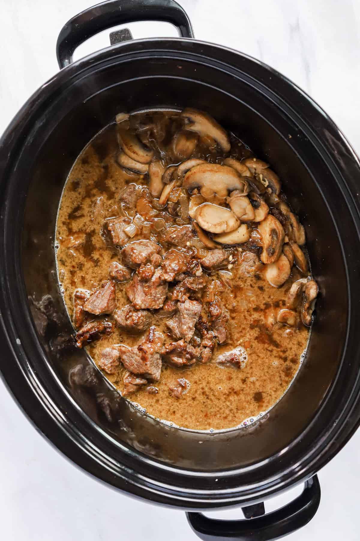 Overhead view inside crockpot of browned beef chunks, mushrooms and broth