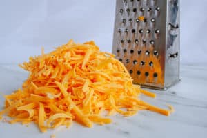 Pile of grated sharp cheddar cheese with metal cheese grater in background.