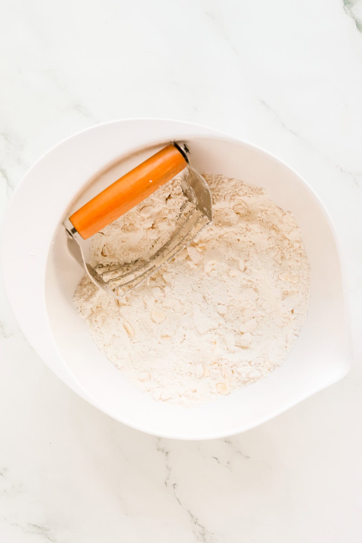Pastry cutter in white mixing bowl with flour and pea-sized butter chunks.