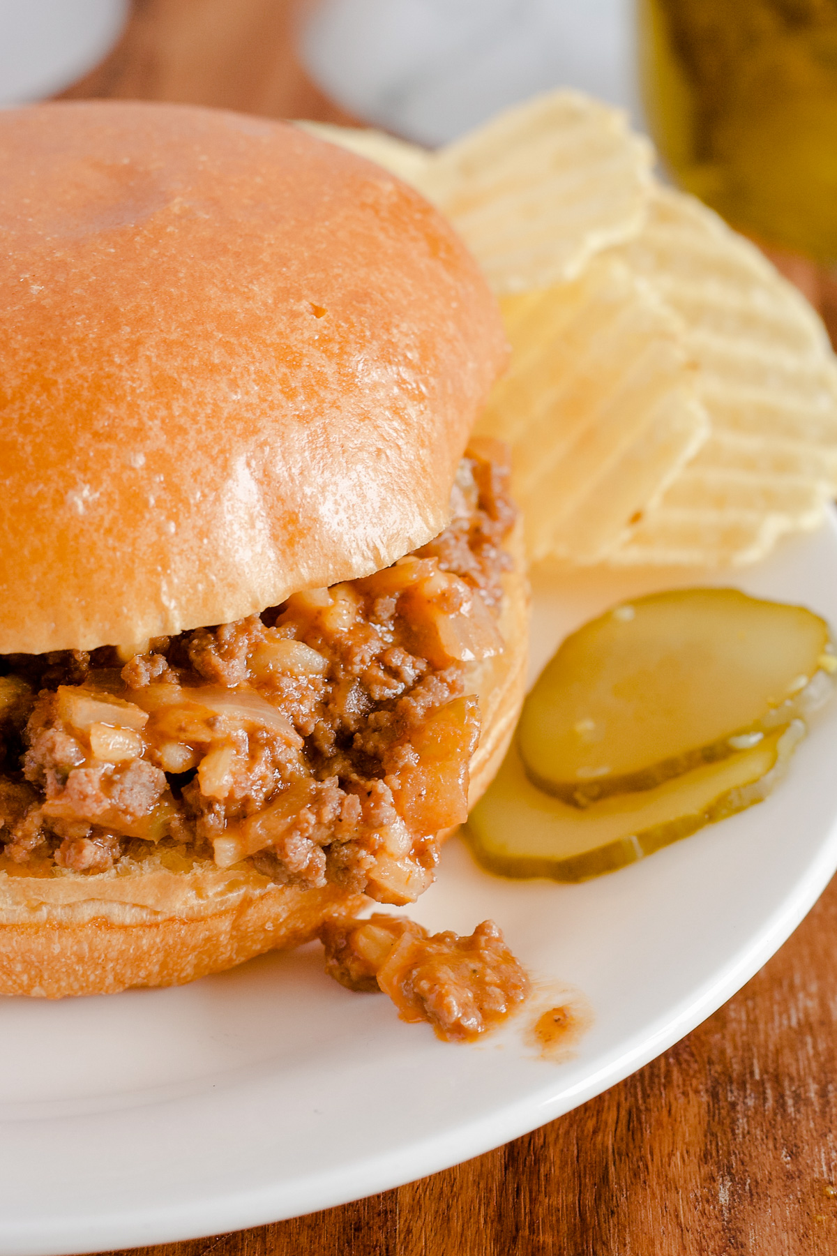 Sloppy Joe with a few pieces of meat falling onto white plate with pickles and chips