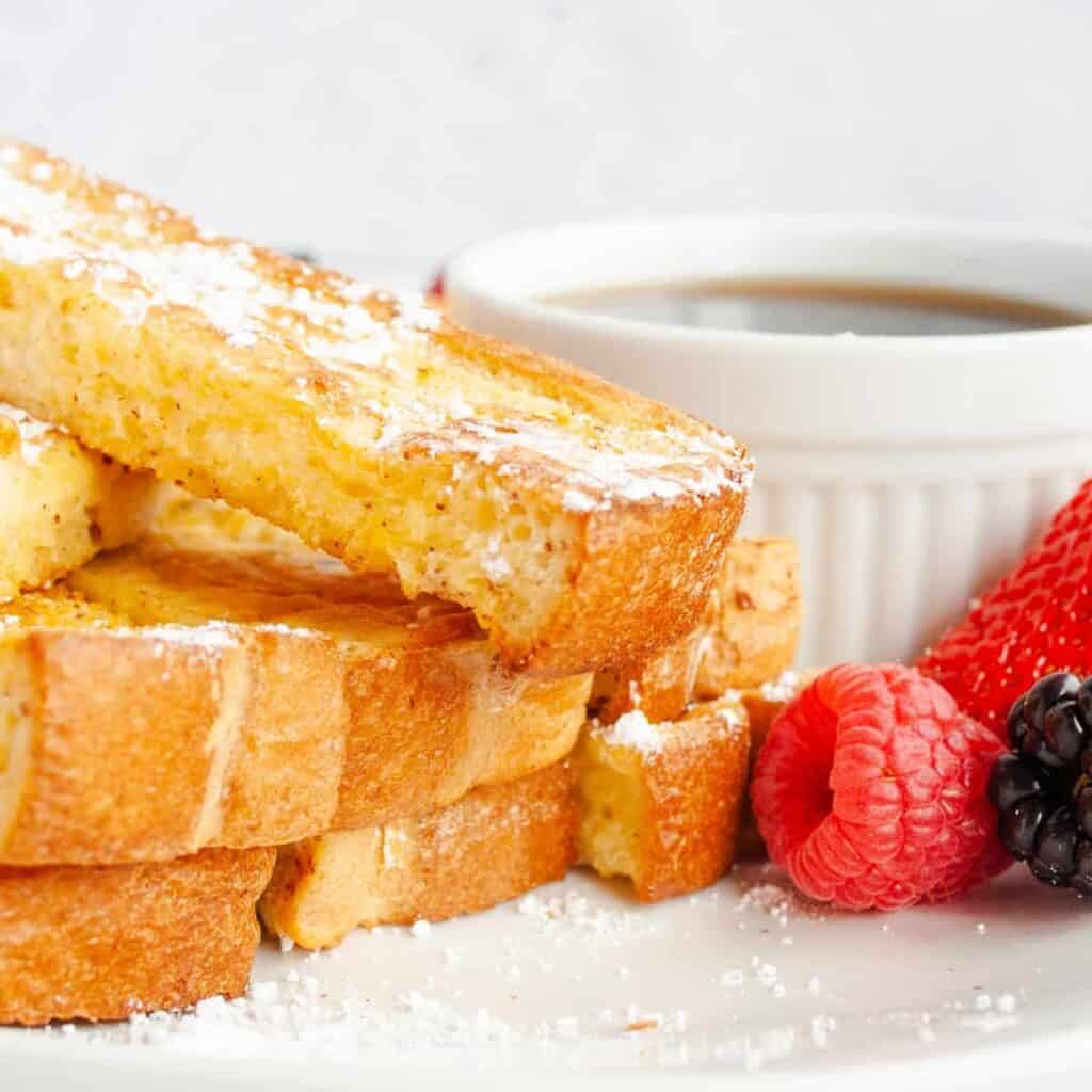 Baked French Toast Sticks on a white plate with a cup of syrup and fresh berries