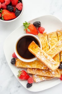 Baked French Toast Sticks on a white plate with a cup of syrup and fresh berries