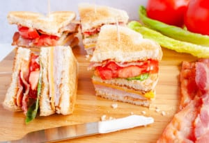 Classic Club Sandwich on wooden cutting board along side lettuce, tomatoes and bacon slices