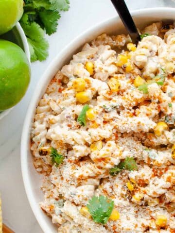 Mexican Street Corn Pasta Salad in a white bowl alongside whole limes and corn on the cob