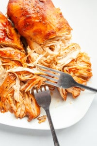 Cooked chicken breasts on a white plate being shredded with two forks 