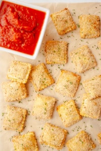 Crispy Baked Ravioli on brown parchment with a white bowl of marinara sauce