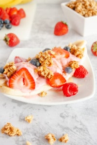 Breakfast Banana Split on a white plate with extra berries and granola on countertop