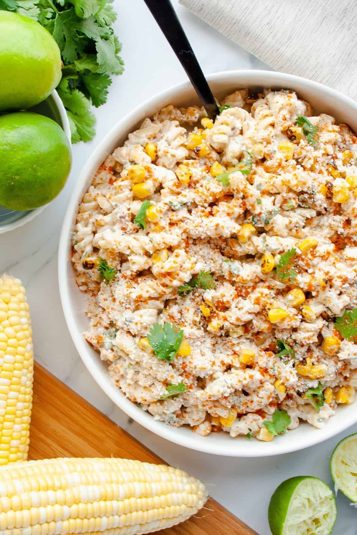 Street corn pasta salad in a serving bowl next to corn cobs, cilantro and limes.