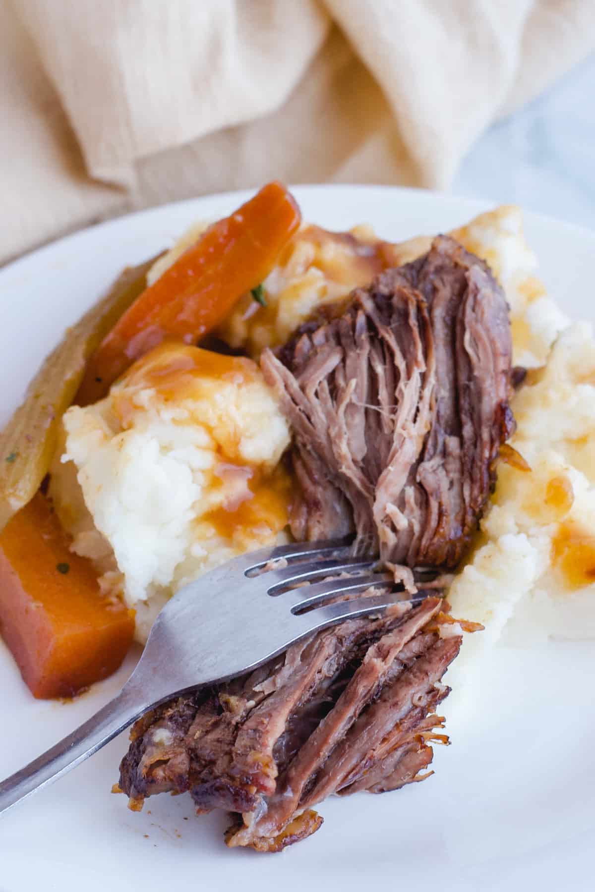 Fork shredding a piece of pot roast on a white plate with mashed potatoes, gravy and vegetables