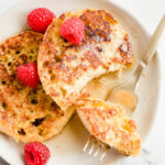 2 pieces of French toast on white plate with syrup and raspberries and a small piece on a fork