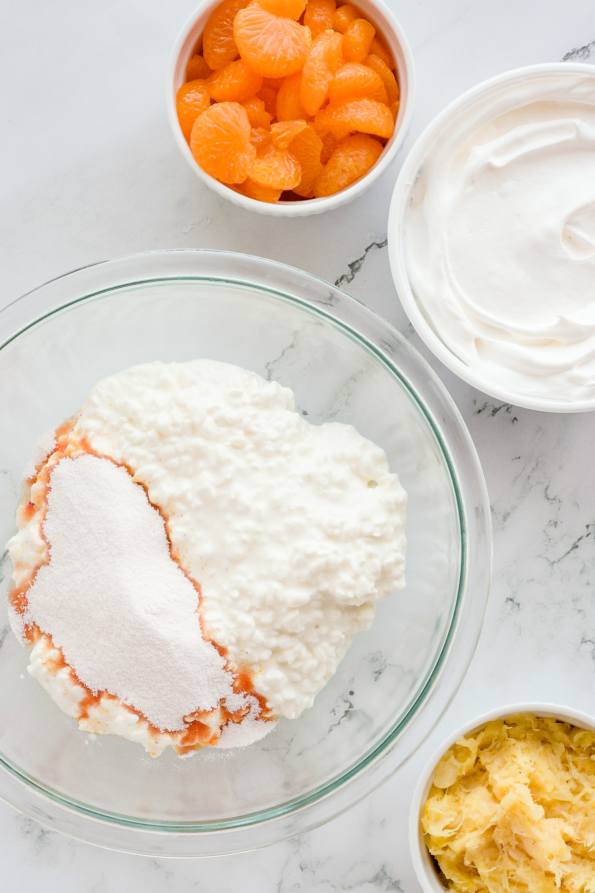 Jello salad ingredients on countertop with orange jello powder sprinkled on top of cottage cheese in glass bowl