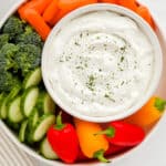 Dip in a white bowl on a round tray with fresh carrots, broccoli, cucumber and mini peppers