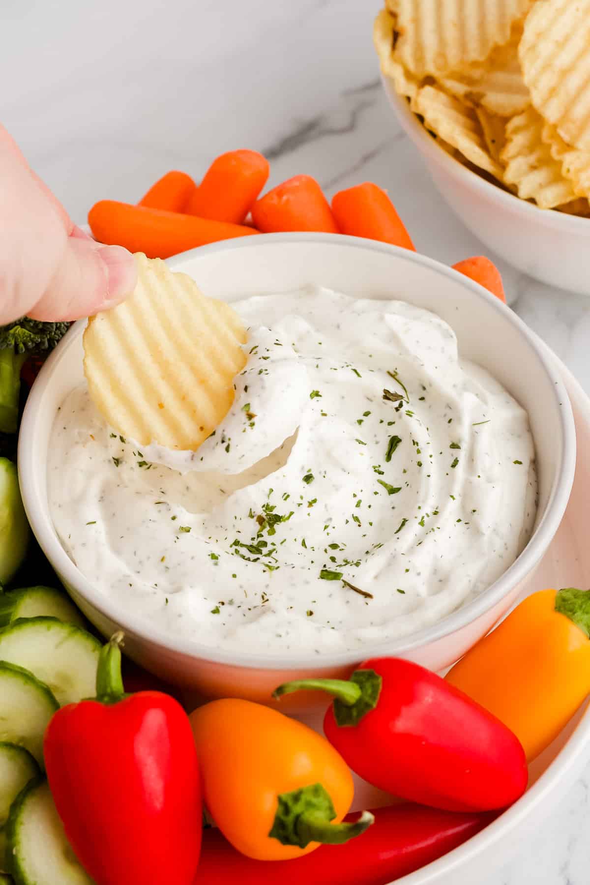 Hand dipping a potato chip into dip bowl surrounded by fresh veggies and bowl of potato chips in the background.