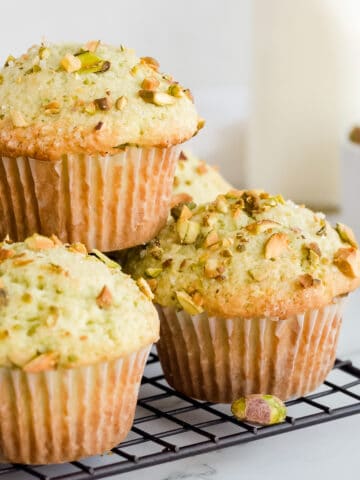Pistachio muffins stacked on wire rack with a glass of milk and a bowl of pistachios in background