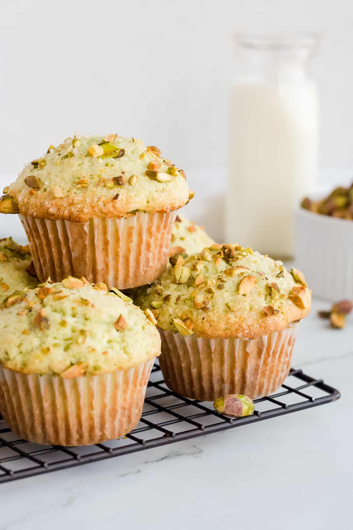 Pistachio muffins stacked on wire rack with a glass of milk and a bowl of pistachios in background