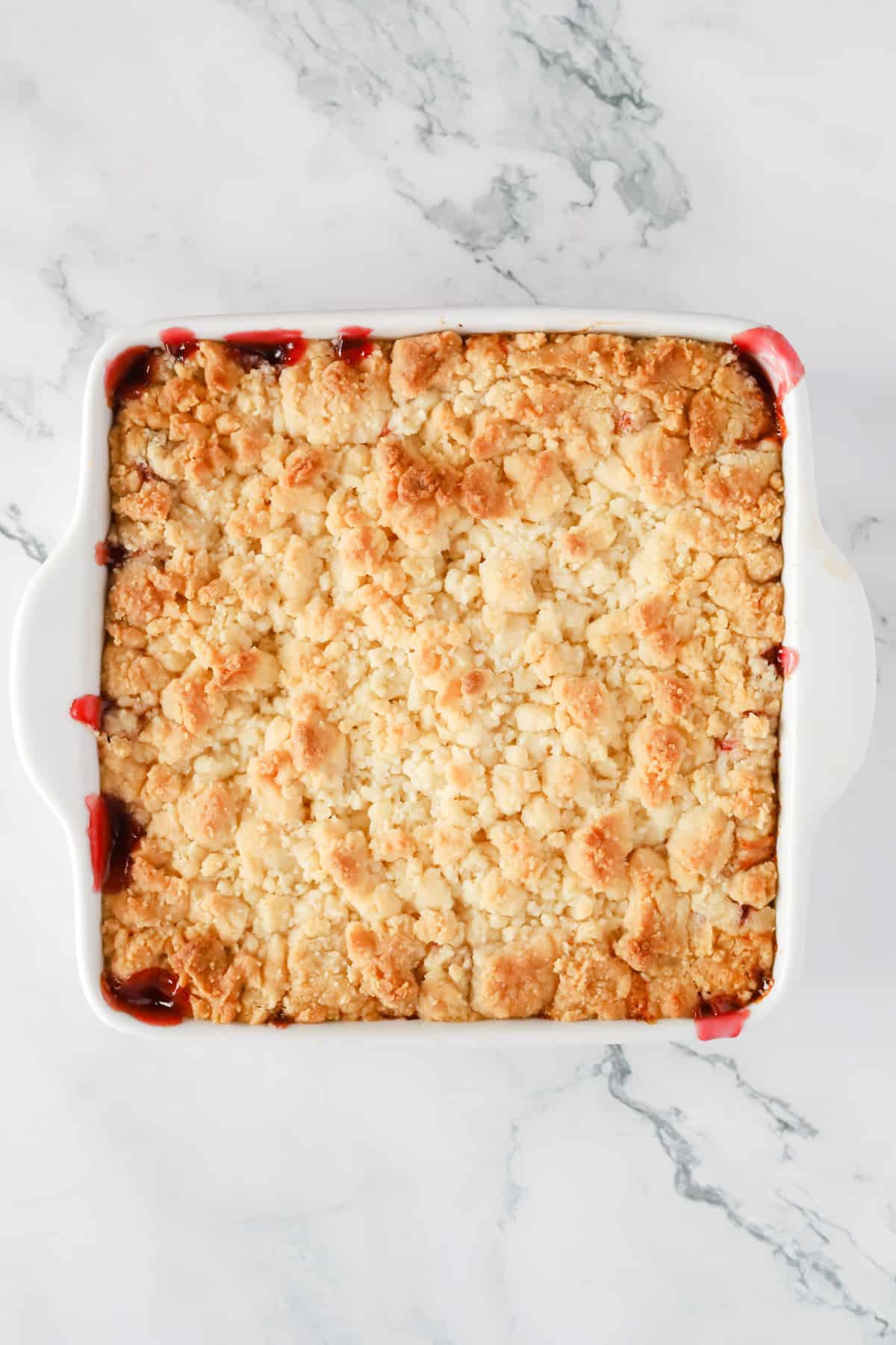 Baked cherry cobbler in a white 8x8 baking dish. The topping is golden brown and some cherry filling is leaking around the edges. 