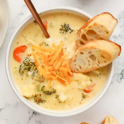 White bowl of cheesy roasted broccoli and cauliflower soup with a copper spoon sticking out. Two slices of toasted baguette are sitting angled in the soup and some extra shredded cheese garnishes the top.