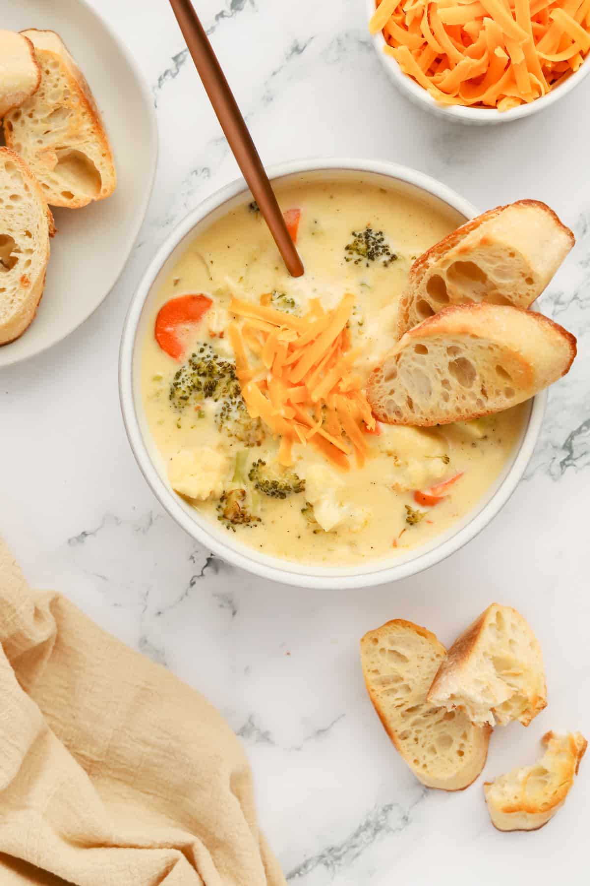 White bowl of soup with 2 pieces of baguette sitting on the side. A white bowl of cheese is on the top right, a plate of toasted baguette on the left and chunks of baguette on the countertop in the bottom right.