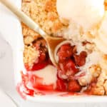 Gold spoon scooping out cherry cobbler. Overhead shot. Scoops of vanilla ice cream are placed on top of the cobbler in the white baking dish.