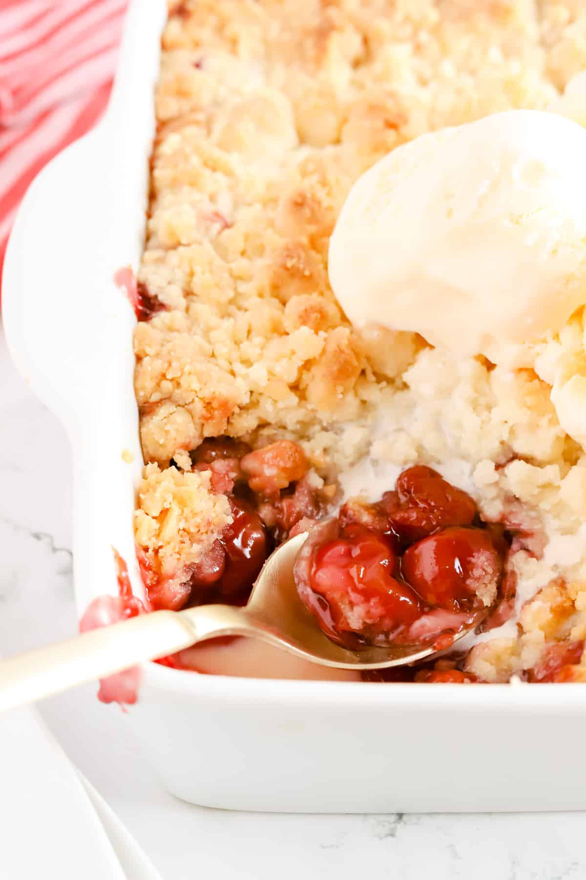 Side view of a gold spoon scooping a helping of cobbler out of a white baking dish. Scoops of vanilla ice cream sit on top of the pan of cobbler.