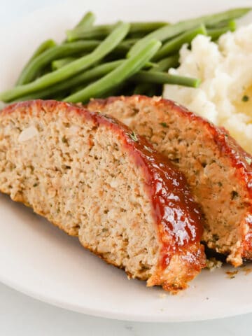 Two slices of meatloaf with mashed potatoes and green beans on a white plate