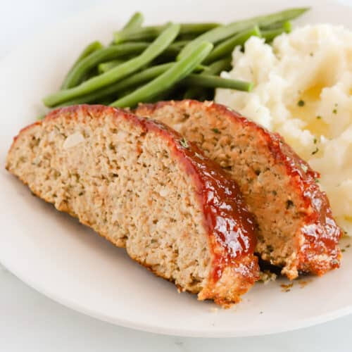 Two slices of meatloaf with mashed potatoes and green beans on a white plate