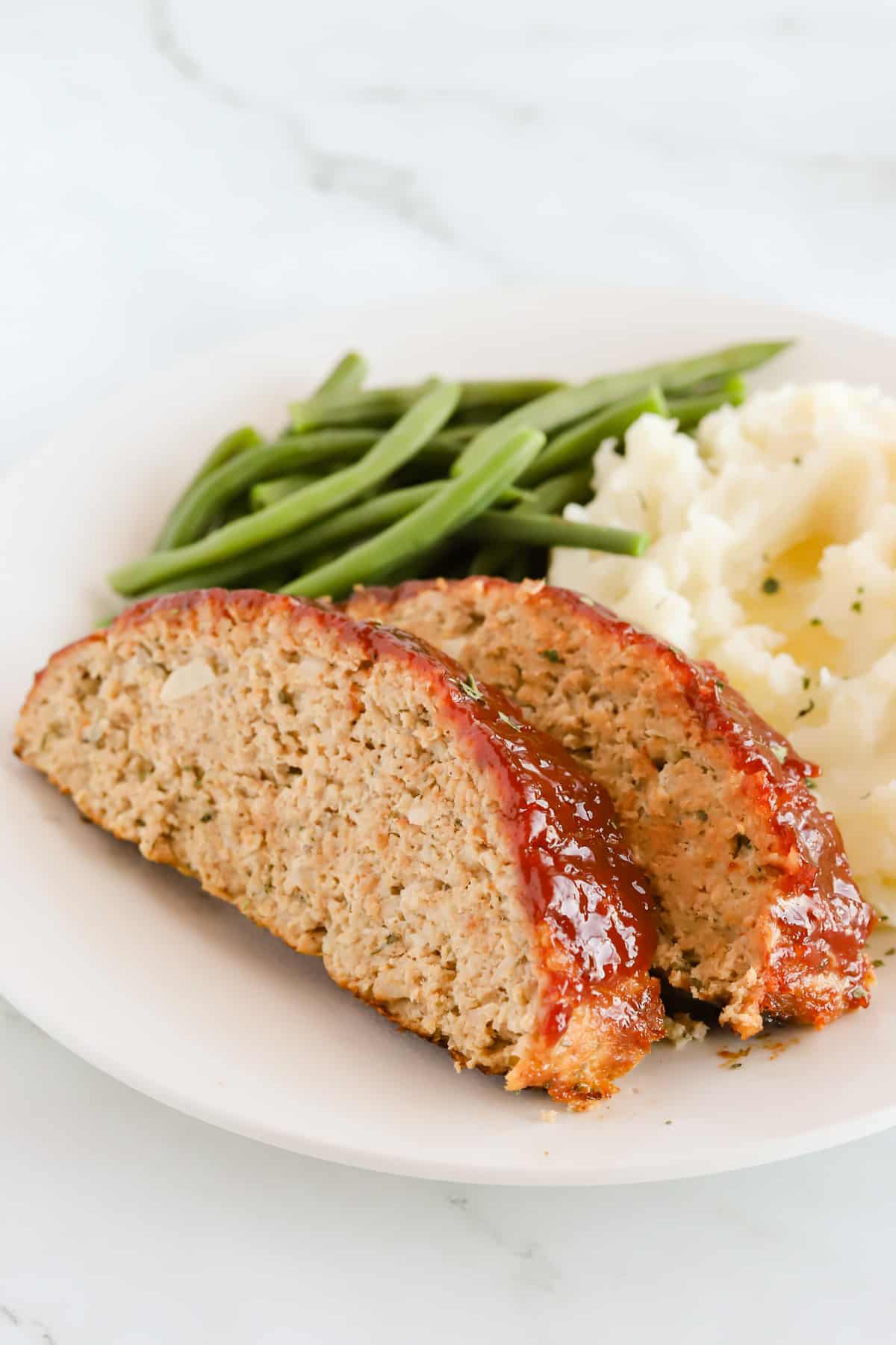 Two slices of meatloaf with mashed potatoes and green beans on a white plate