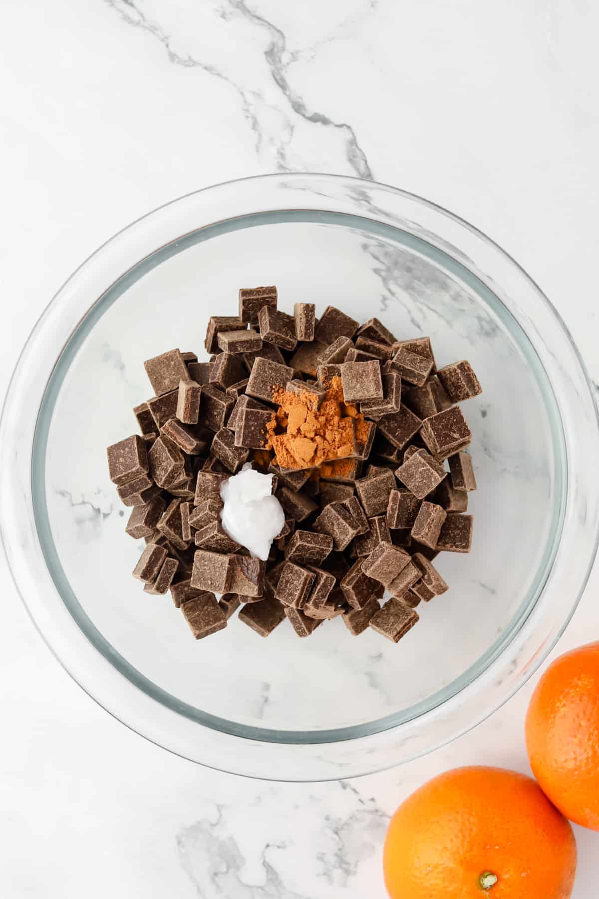 Dark chocolate, coconut oil and cinnamon in a glass bowl alongside two mandarin oranges on marble countertop