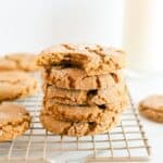 Stack of molasses cookies with a bite taken out of the top cookie all on a gold cooling rack with other cookies surrounding. A glass of milk is in the background.