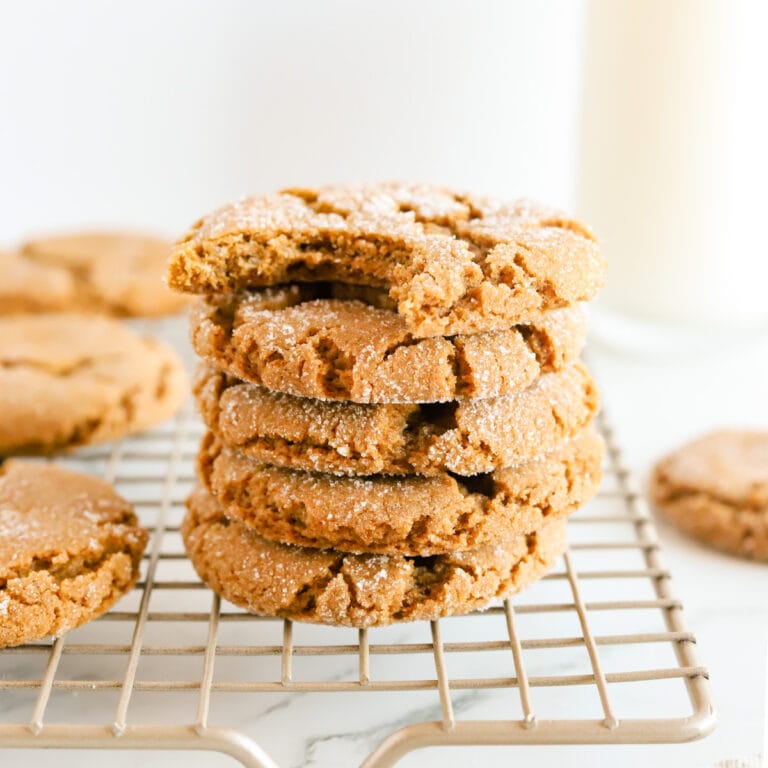 Stack of molasses cookies with a bite taken out of the top cookie all on a gold cooling rack with other cookies surrounding. A glass of milk is in the background.