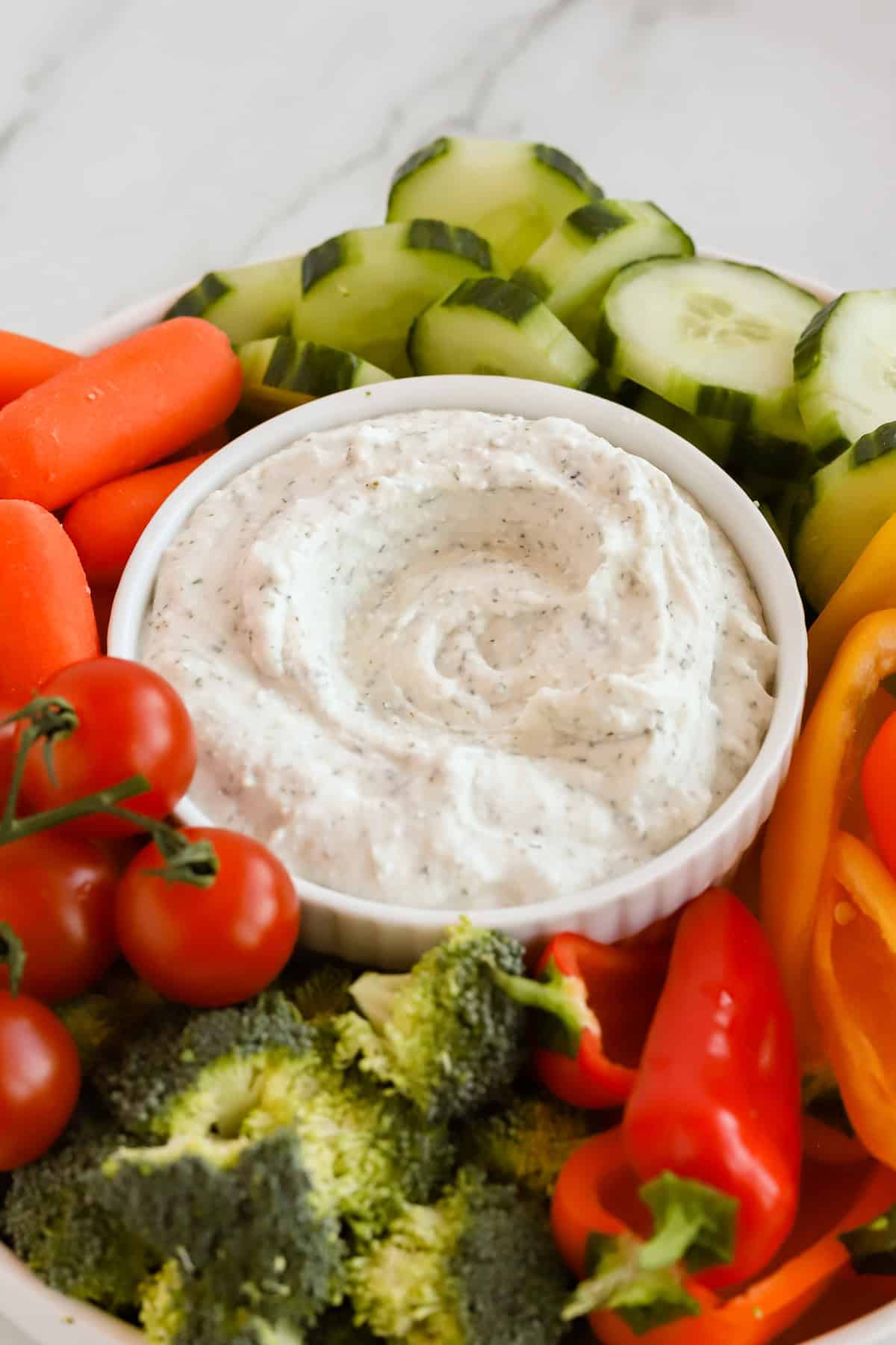 Ranch dip in white bowl surrounded by fresh cucumbers, peppers, carrots, tomatoes and broccoli.