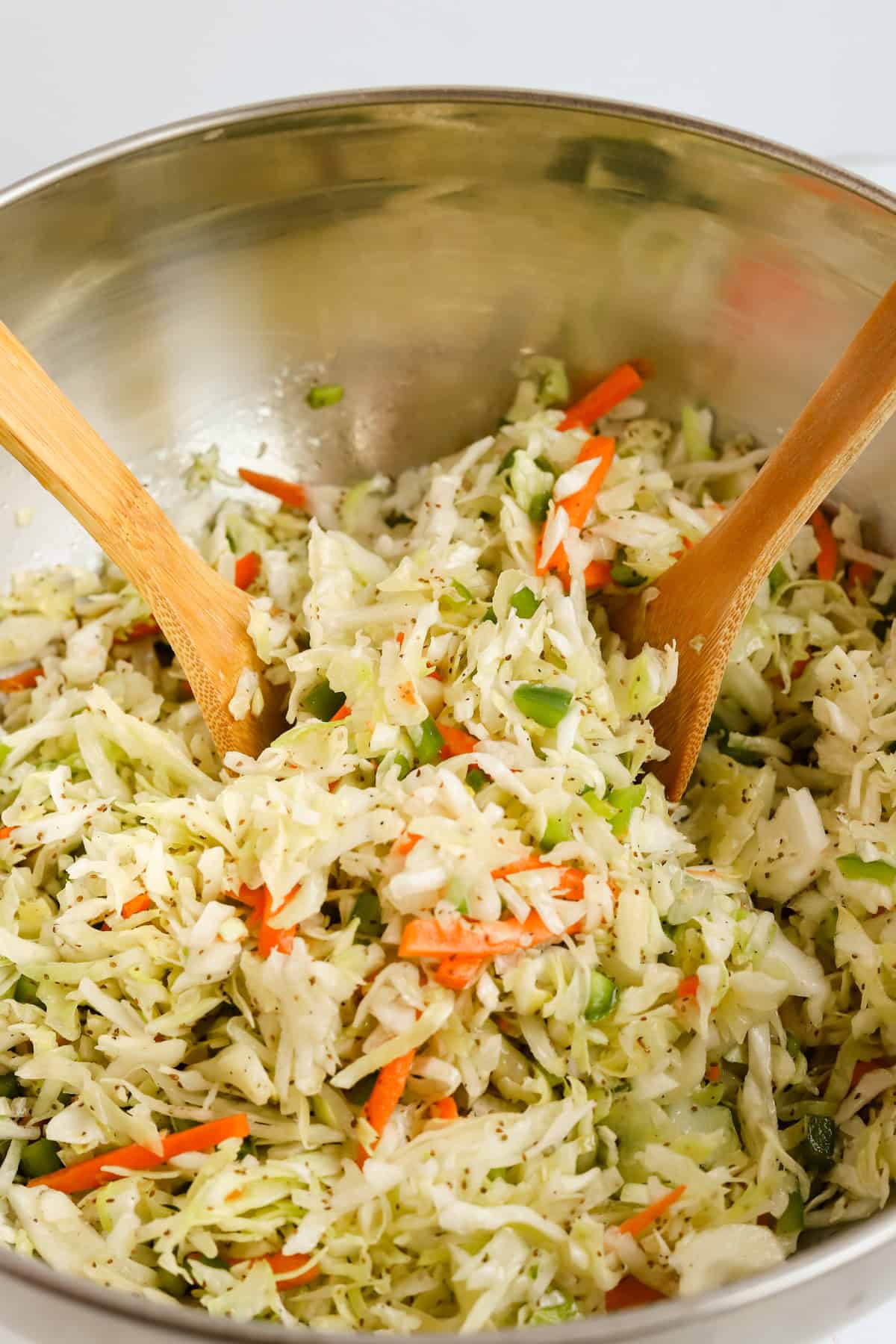 Two wooden spoons scooping coleslaw out of a large metal mixing bowl.
