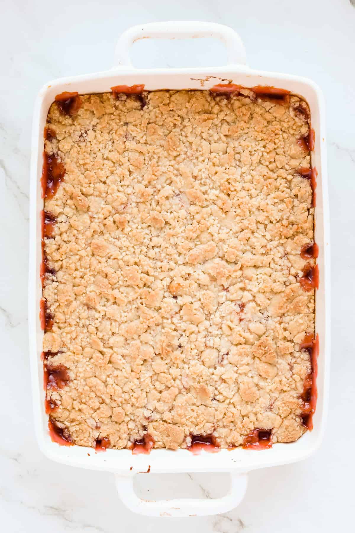 Baked cherry cobbler in a white 8x8 baking dish. The topping is golden brown and some cherry filling is leaking around the edges.