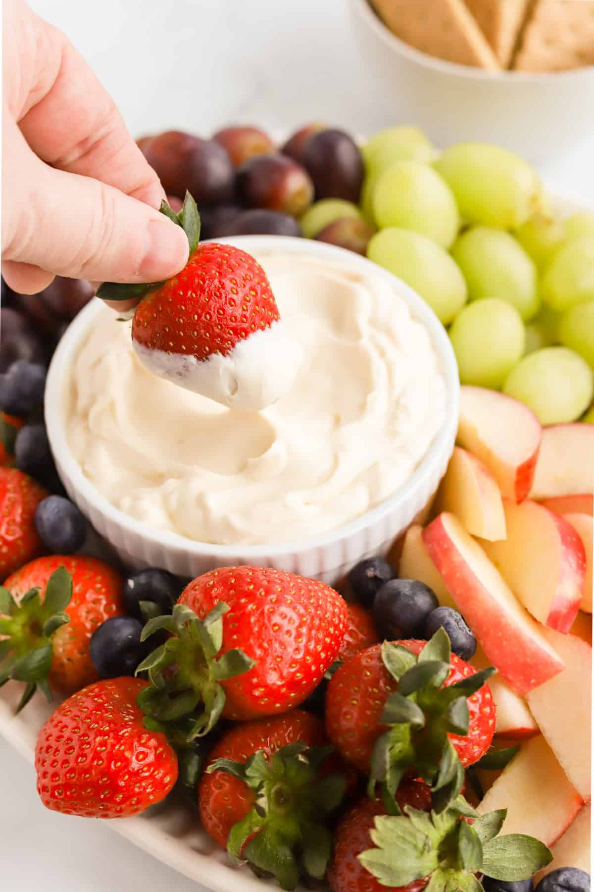 Hand dipping a whole strawberry into mascarpone dip surrounded by fresh fruit and a bowl of graham crackers in the background.