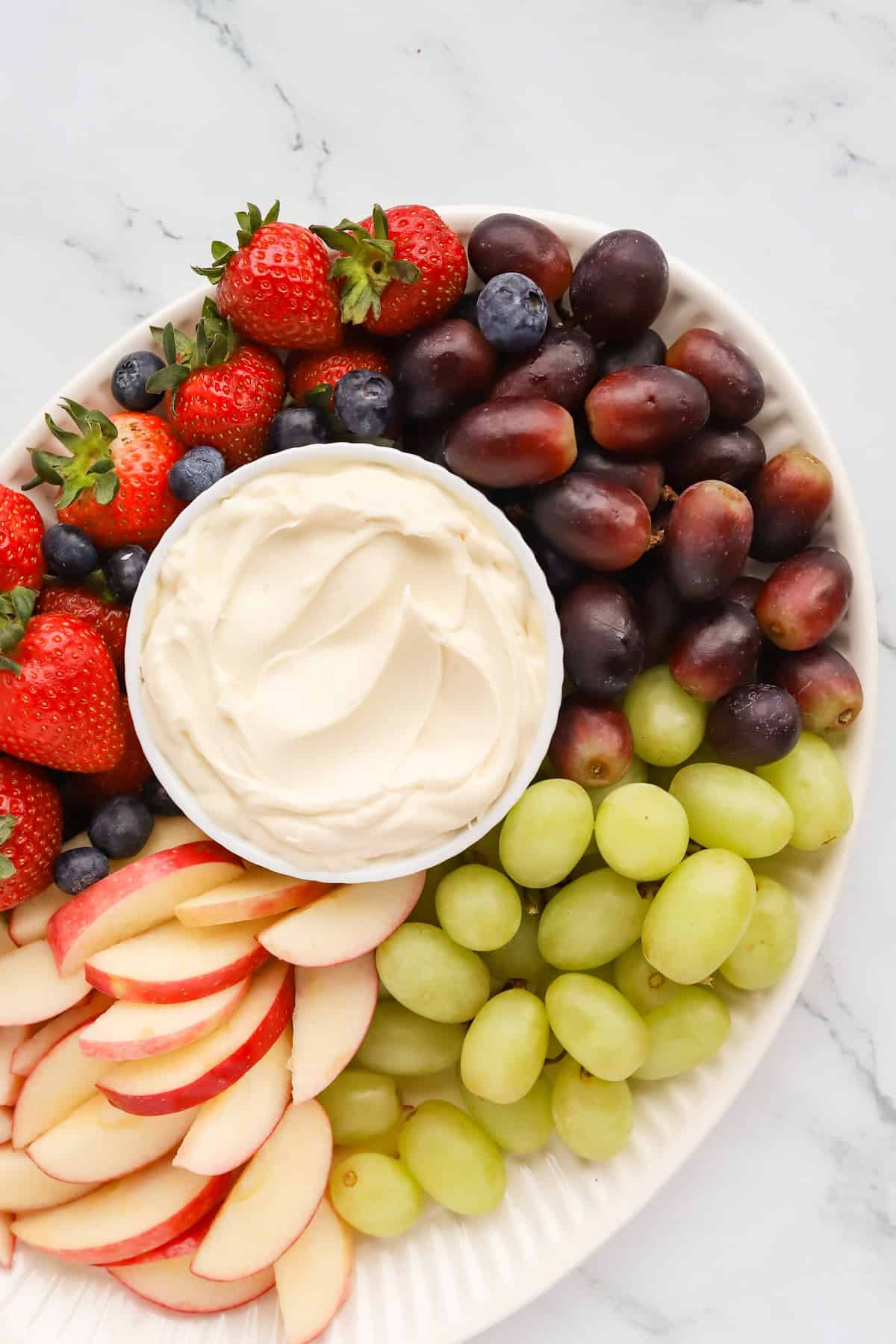 Oval white platter of fresh red and green grapes, strawberries, sliced apples and blueberries with a small white bowl of dip in the middle. 