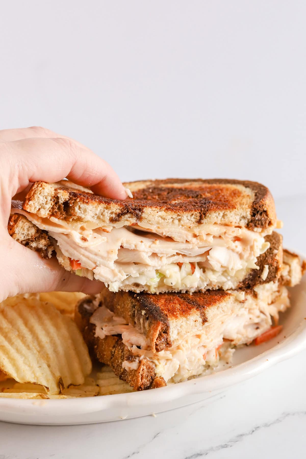 Hand grabbing half of a Rachel sandwich that's sitting on top of other half on a white plate with ruffled potato chips.