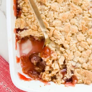Gold spoon scooping out cherry cobbler that's in a white baking dish with a red napkin underneath the corner.