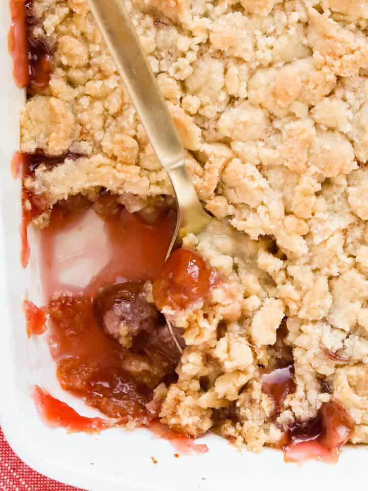 Gold spoon scooping out cherry cobbler that's in a white baking dish with a red napkin underneath the corner.