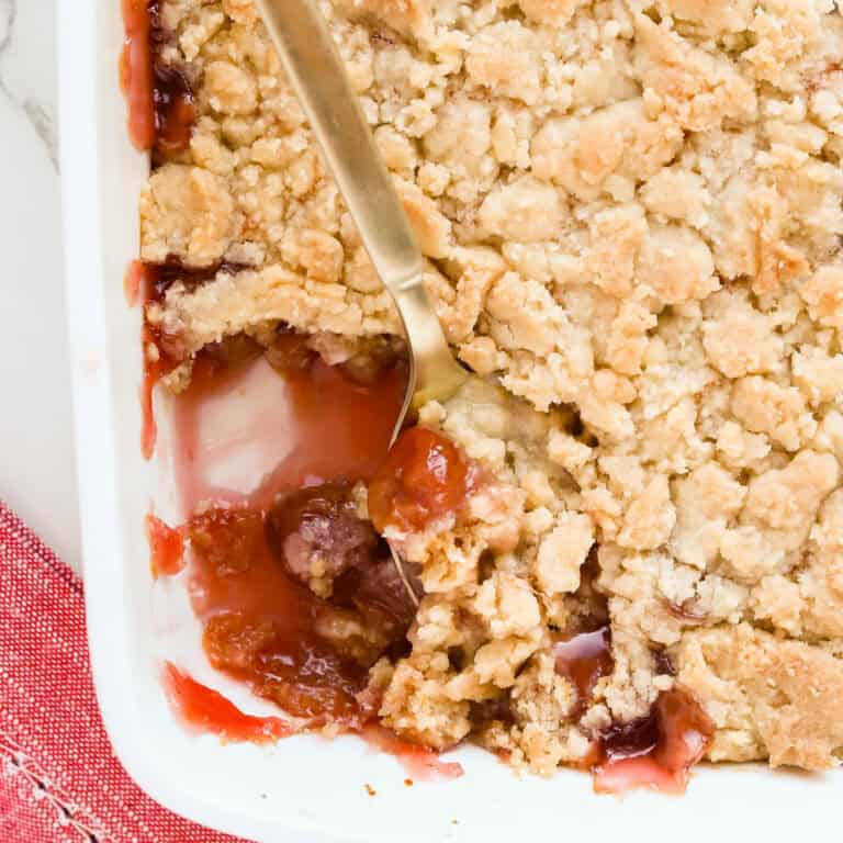 Gold spoon scooping out cherry cobbler that's in a white baking dish with a red napkin underneath the corner.