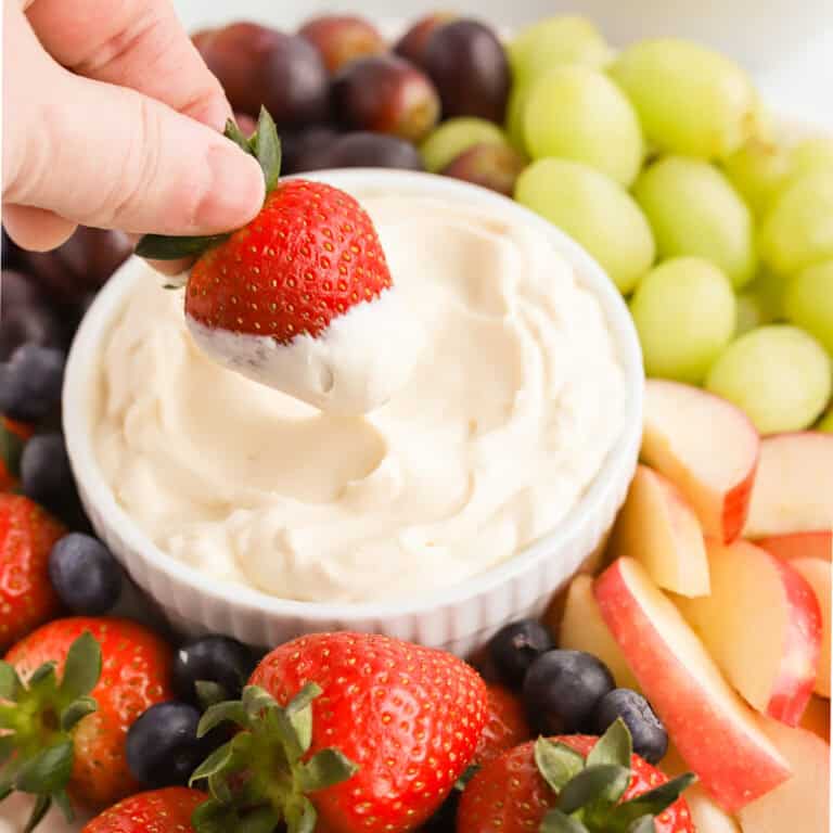Hand dipping a whole strawberry into mascarpone dip surrounded by fresh fruit and a bowl of graham crackers in the background.