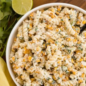 Overhead view of pasta salad in a white bowl sitting on a wooden tray. A yellow towel in the bottom left corner and cilantro and half a lime in the top left corner.