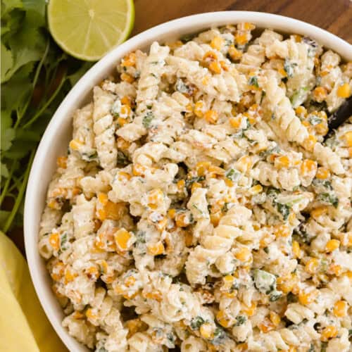 Overhead view of pasta salad in a white bowl sitting on a wooden tray. A yellow towel in the bottom left corner and cilantro and half a lime in the top left corner.