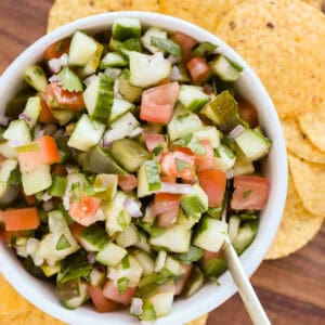 White bowl of pickle salsa with a gold spoon resting in it on a wooden tray with yellow corn tortilla chips surrounding the bowl.