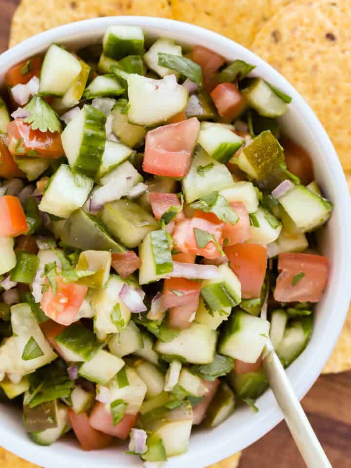 White bowl of pickle salsa with a gold spoon resting in it on a wooden tray with yellow corn tortilla chips surrounding the bowl.