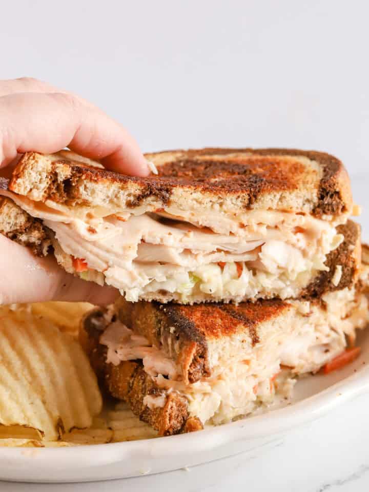 Hand grabbing half of a Rachel sandwich that's sitting on top of other half on a white plate with ruffled potato chips.