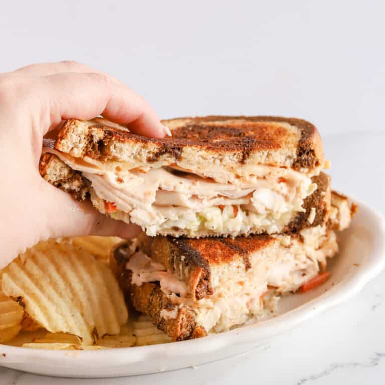 Hand grabbing half of a Rachel sandwich that's sitting on top of other half on a white plate with ruffled potato chips.